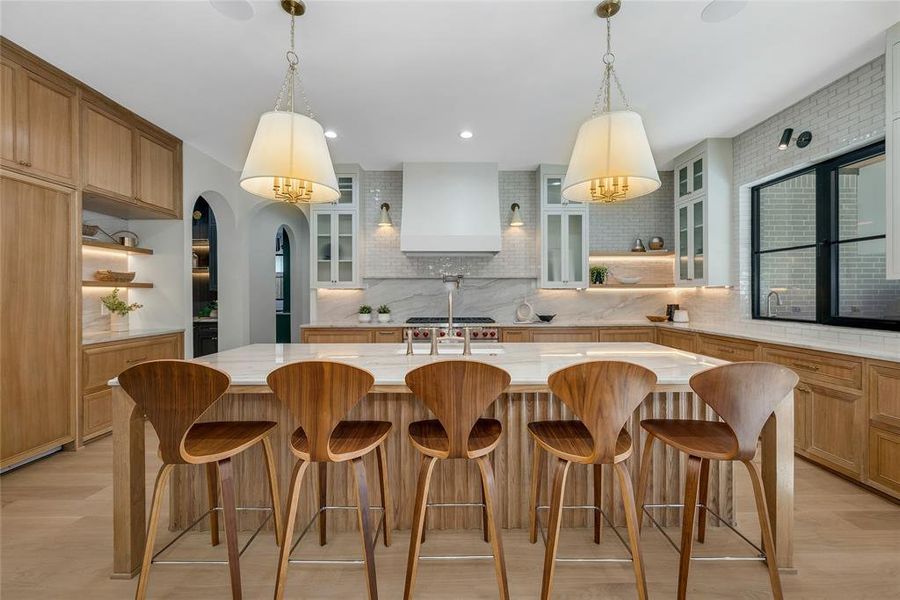 Gourmet kitchen featuring a large central island with seating for five, natural wood-finish cabinetry, quartz countertops, a tile backsplash, and a range hood