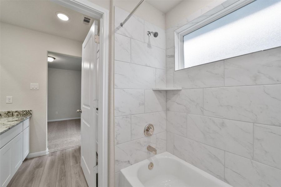 Modern bathroom featuring a tiled shower/tub combo and frosted window, conveniently located near a granite-topped vanity and light wood flooring.