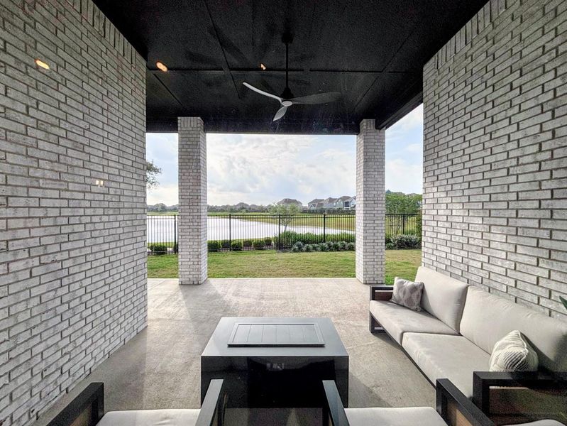 The spacious covered patio features modern outdoor seating, a ceiling fan with large windows offering scenic views of a nearby lake. Perfect for relaxing and entertaining.