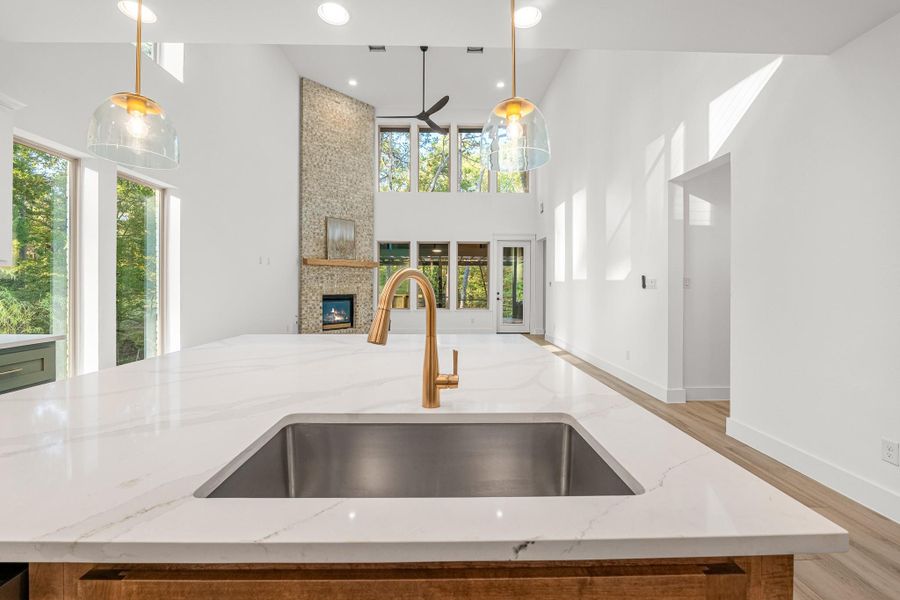 This spacious, modern kitchen features a large quartz island with sleek gold fixtures, high ceilings, and ample natural light. The open layout connects to a living area with a tall stone fireplace and large windows, offering a bright and inviting atmosphere. This spacious, modern kitchen features a large quartz island with sleek gold fixtures, high ceilings, and ample natural light. The open layout connects to a living area with a tall stone fireplace and large windows, offering a bright and inviting atmosphere.