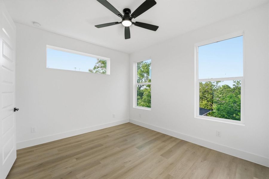 Bright and inviting bedroom featuring wood-look flooring, modern ceiling fan, and multiple windows that provide abundant natural light with peaceful tree-top views.