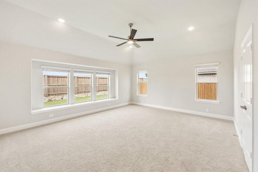 Spacious empty bedroom with vaulted ceiling, ceiling fan, carpet flooring, and multiple windows overlooking a fenced backyard.