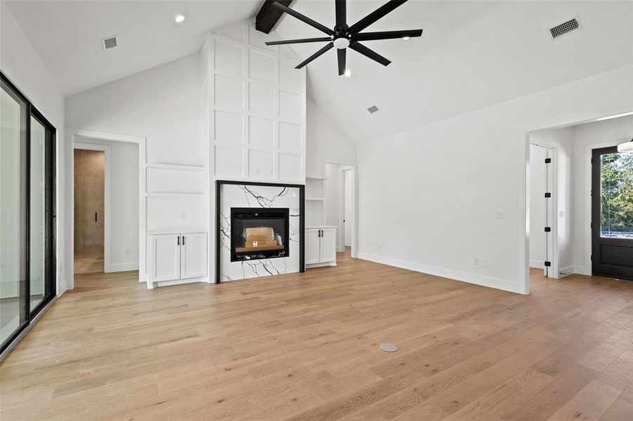 Unfurnished living room with a ceiling fan, light wood finished floors, high vaulted ceiling, beam ceiling, and a high end fireplace Unfurnished living room with a ceiling fan, light wood finished floors, high vaulted ceiling, beam ceiling, and a high end fireplace
