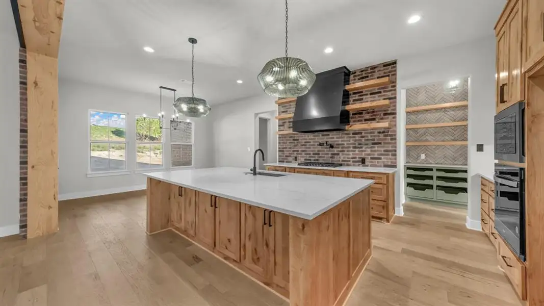 Spacious kitchen featuring a large central island with a solid surface countertop, wood-finish cabinetry, a brick-textured accent wall with open shelving, and built-in appliances Spacious kitchen featuring a large central island with a solid surface countertop, wood-finish cabinetry, a brick-textured accent wall with open shelving, and built-in appliances