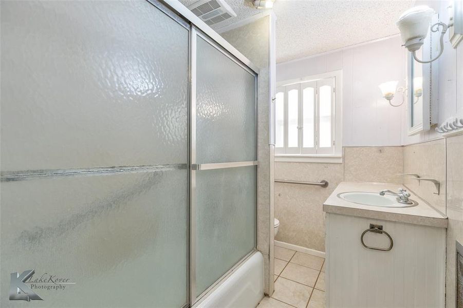 Full bath featuring vanity, combined bath / shower with glass door, light tile patterned floors, and a textured ceiling Full bath featuring vanity, combined bath / shower with glass door, light tile patterned floors, and a textured ceiling