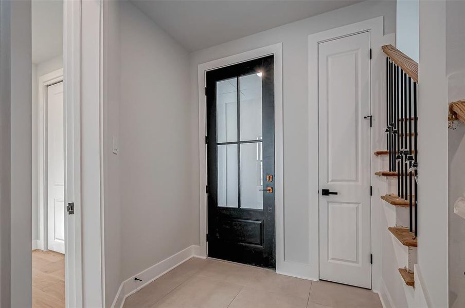 This photo shows a modern entryway with a sleek black front door featuring glass panels, allowing natural light to enter. Adjacent is a white door opening to a storage area under stairs, and a staircase with wooden steps and black metal railings. The space has light-color walls and flooring, creating a bright and welcoming ambiance.