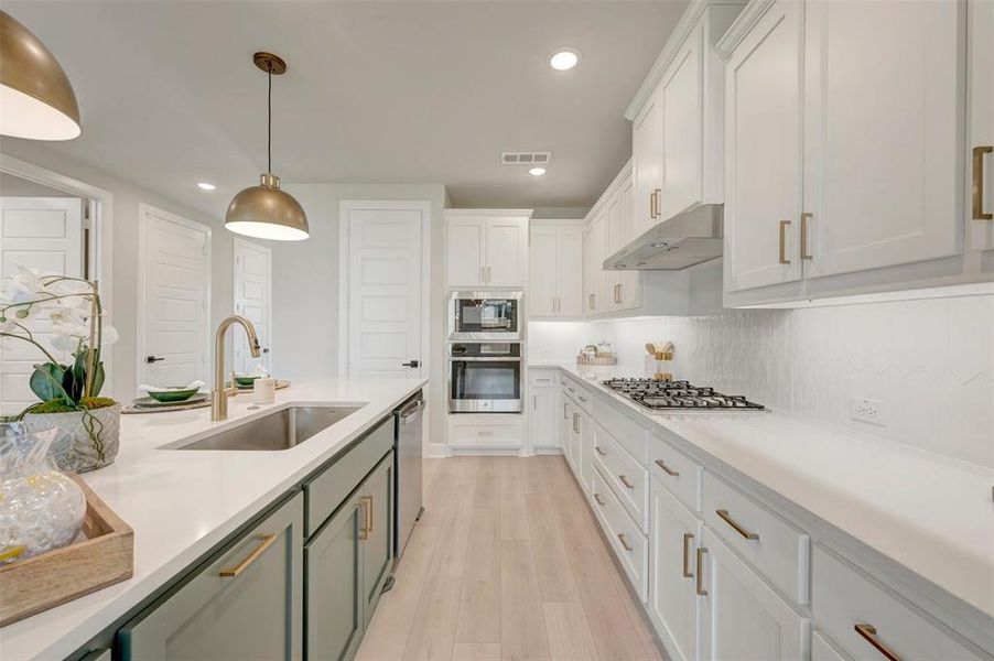Kitchen featuring a central island with an undermount sink and brushed brass faucet, white cabinetry, integrated stainless steel appliances, light wood-finish flooring, and recessed lighting
