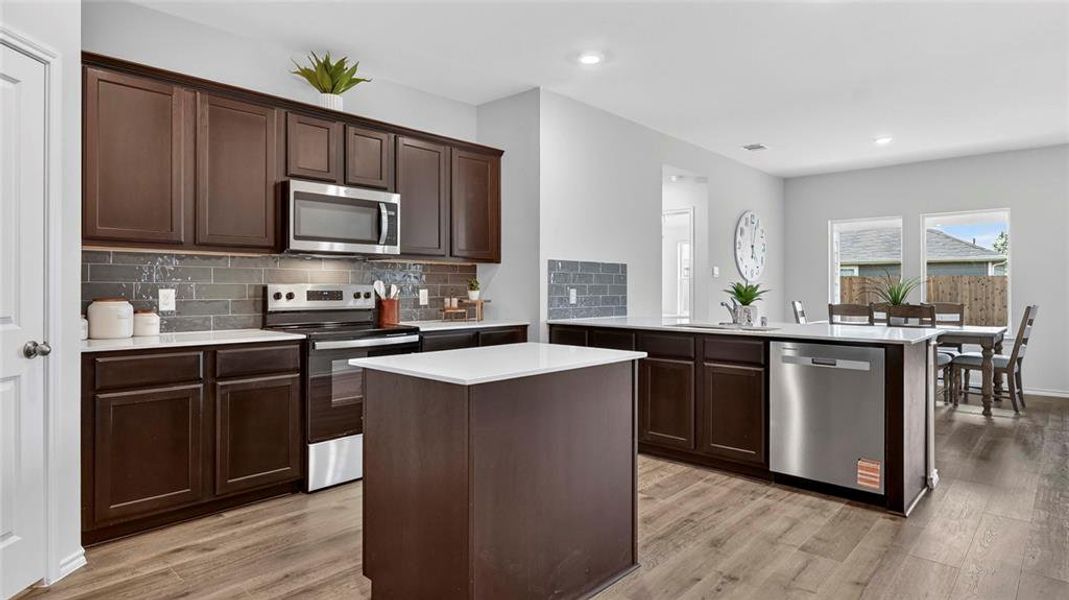 Kitchen featuring dark wood cabinetry, stainless steel appliances, a center island with a white countertop, and wood-finish flooring Kitchen featuring dark wood cabinetry, stainless steel appliances, a center island with a white countertop, and wood-finish flooring