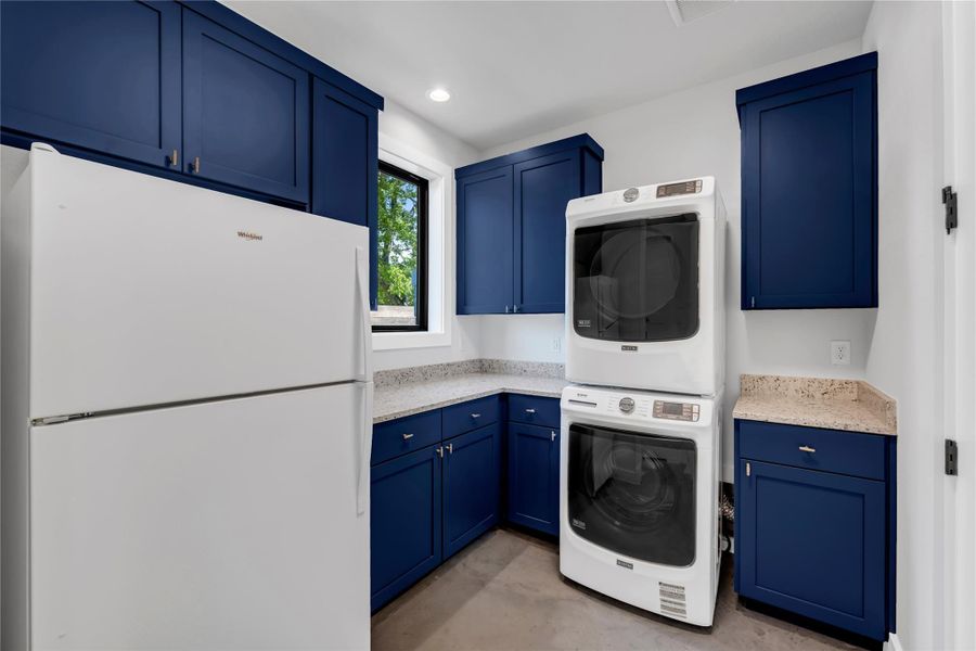 Laundry area featuring stacked washer and dryer, blue shaker-style cabinetry, light-colored speckled countertops, and a white refrigerator