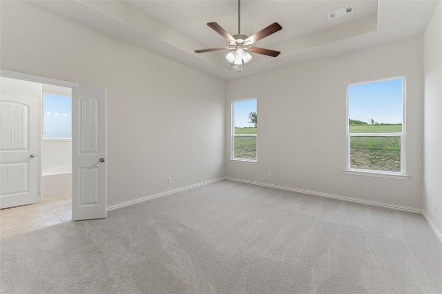 Spacious interior room with light gray wall paint, gray carpet flooring, and a tray ceiling with a ceiling fan light fixture