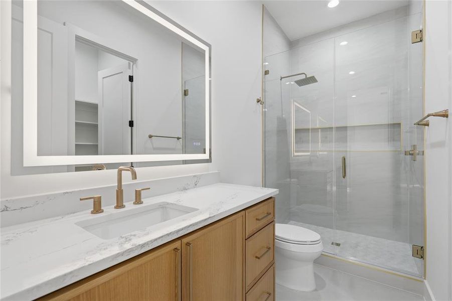 Bathroom featuring a large vanity with an integrated sink and light wood-finish cabinetry, a lighted mirror, a glass-enclosed shower with a rain showerhead, and gold-tone fixtures Bathroom featuring a large vanity with an integrated sink and light wood-finish cabinetry, a lighted mirror, a glass-enclosed shower with a rain showerhead, and gold-tone fixtures