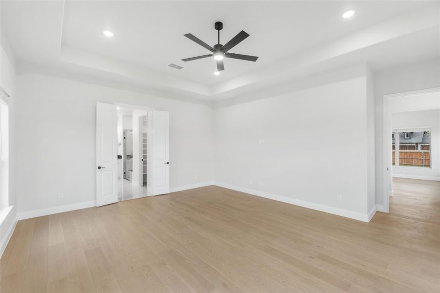 Empty bedroom with tray ceiling, black ceiling fan, light hardwood floors, and double doors leading to ensuite bathroom