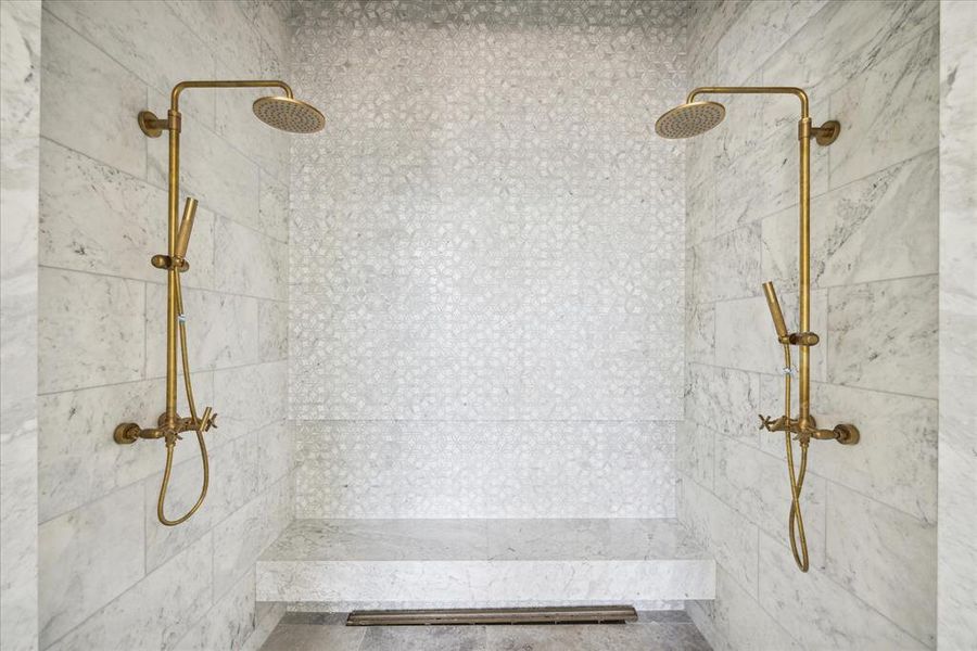 View of the beautiful primary shower situated with a marble bench, two gold rainfall shower heads with handhelds.