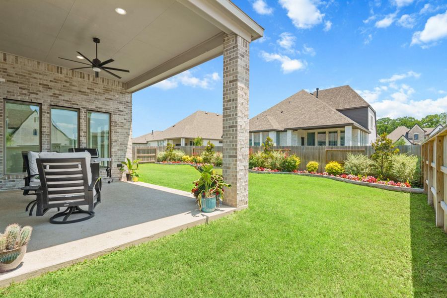 This photo showcases a well-maintained backyard with a covered patio featuring a ceiling fan, seating area, and a brick exterior. The lush lawn is bordered by colorful flowerbeds, offering a serene outdoor space ideal for relaxation or entertaining.