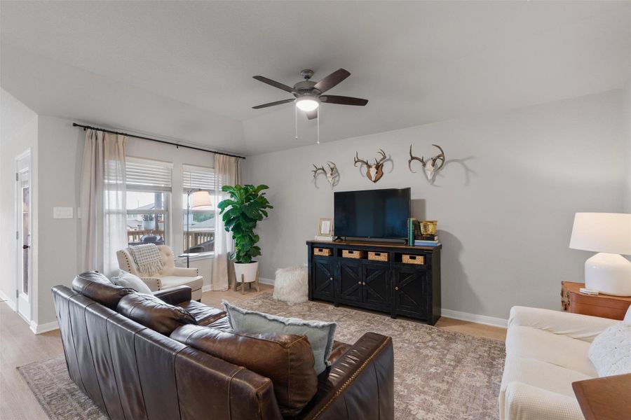 Living area featuring light gray walls, wood-finish flooring, a ceiling fan, and large windows with blinds and curtains Living area featuring light gray walls, wood-finish flooring, a ceiling fan, and large windows with blinds and curtains