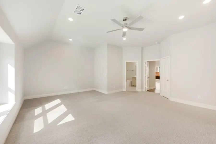 Spacious empty bedroom with vaulted ceiling, ceiling fan, carpet flooring, and ensuite bathroom doorway