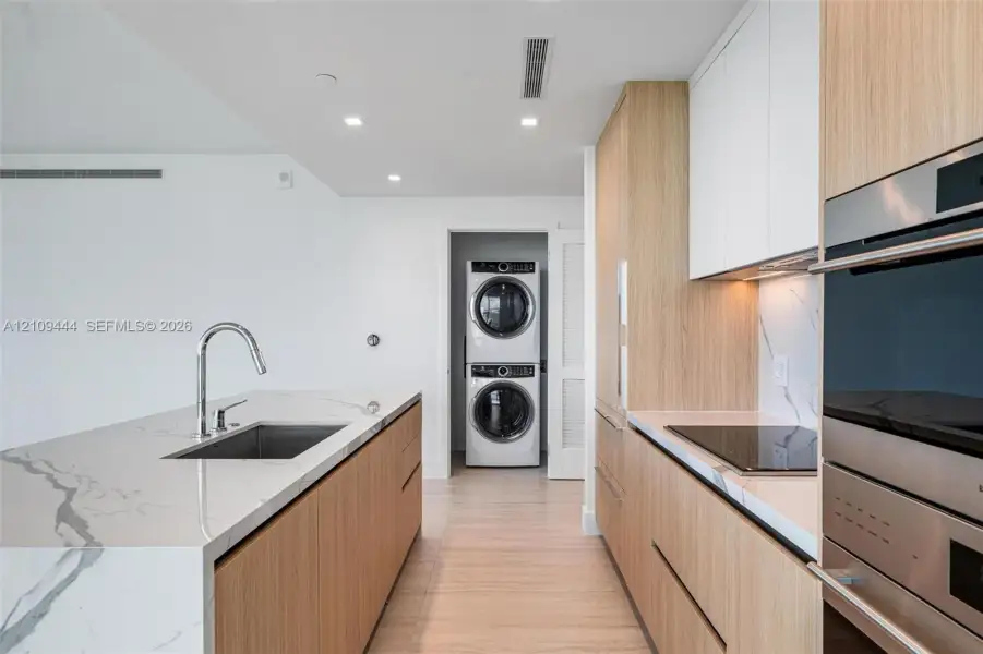 Kitchen featuring wood-finish cabinetry, stone-surface countertops, stainless steel undermount sink with gooseneck faucet, built-in wall oven, and stacked laundry unit
