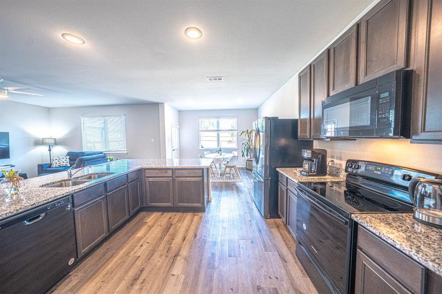 Kitchen with dark brown cabinetry, black appliances, open floor plan, light wood-type flooring, and light stone counters