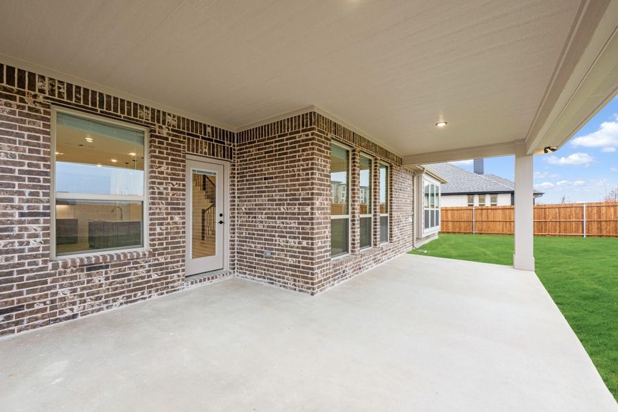 Covered concrete patio with brick exterior, large windows, and fenced backyard with green lawn Covered concrete patio with brick exterior, large windows, and fenced backyard with green lawn
