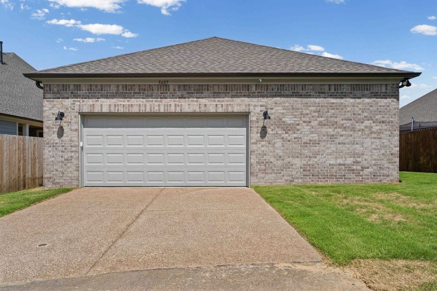 Front exterior featuring a paneled garage door, light-colored brick facade, dark roofing, and a concrete driveway Front exterior featuring a paneled garage door, light-colored brick facade, dark roofing, and a concrete driveway