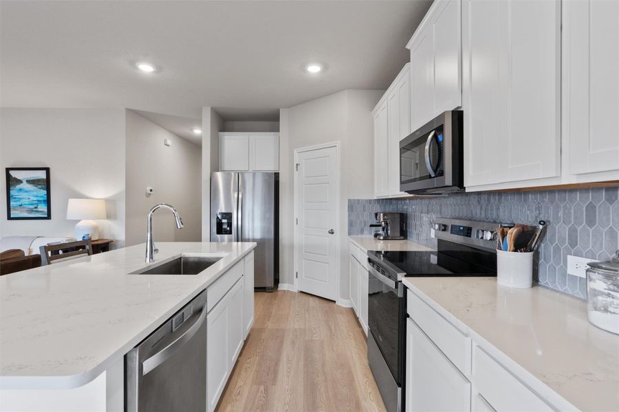 Modern kitchen featuring an island with an undermount sink, stainless steel appliances, white cabinetry, wood-finish flooring, and a unique tiled backsplash Modern kitchen featuring an island with an undermount sink, stainless steel appliances, white cabinetry, wood-finish flooring, and a unique tiled backsplash