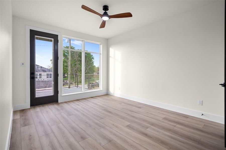 This room features light wood-look flooring, a modern ceiling fan with wooden blades, and a large window providing natural light This room features light wood-look flooring, a modern ceiling fan with wooden blades, and a large window providing natural light