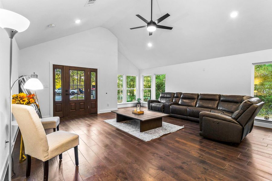 This spacious living room features high vaulted ceilings, hardwood floors, and abundant natural light from large windows. It includes a modern ceiling fan, a comfortable leather sectional, and a cozy seating area, creating an inviting and stylish space for relaxation.