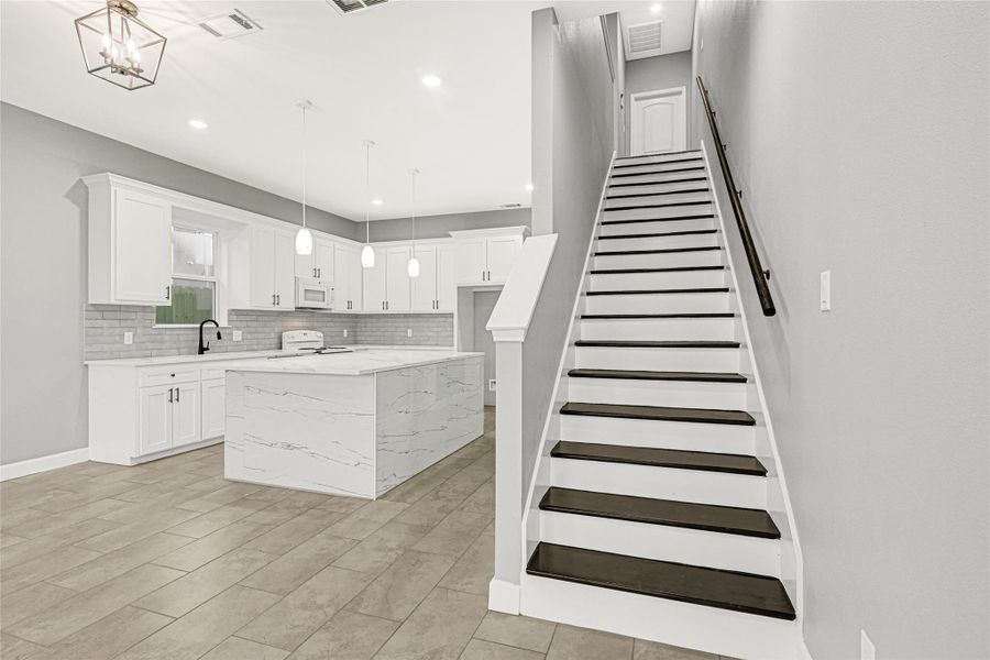 This photo showcases a modern, open-concept kitchen and stairway. The kitchen features sleek white cabinets, a large marble island, and pendant lighting. The space is bright and inviting, with neutral tones and a clean, contemporary design.