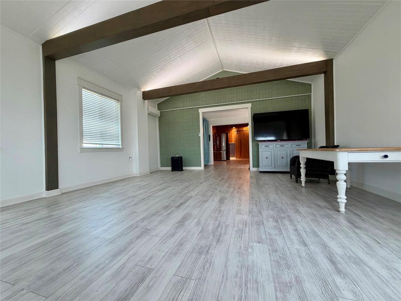 A spacious room features vaulted ceilings, exposed beams, and light-toned wood flooring.