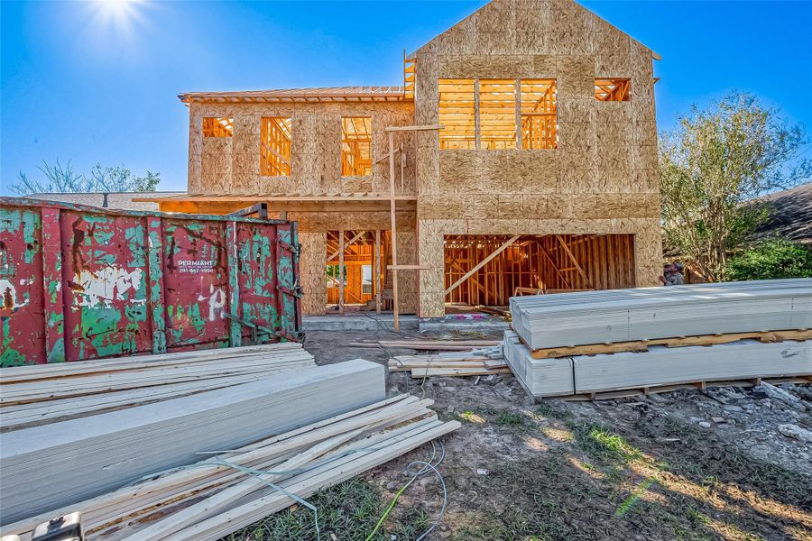 The photo shows a two-story house under construction with exposed wooden framing. Building materials and a large dumpster are visible in the foreground, indicating ongoing work.
