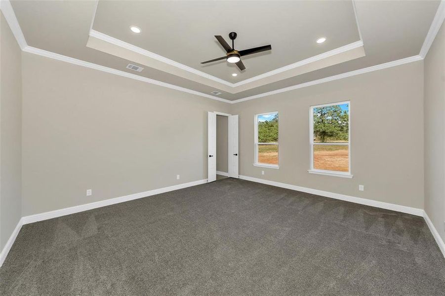 Empty room with a tray ceiling, dark colored carpet, ceiling fan, ornamental molding, and recessed lighting