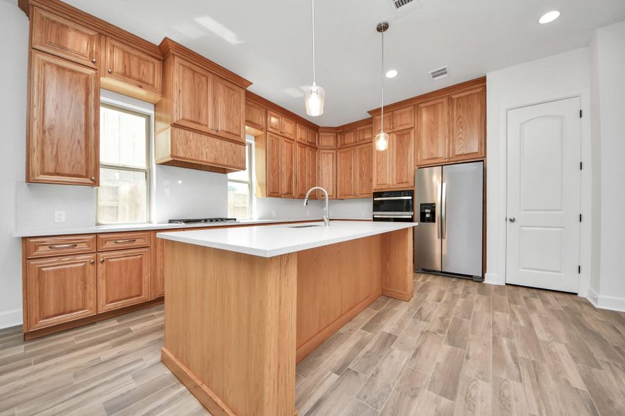 This kitchen features warm wood cabinetry, a large island with a modern sink, stainless steel appliances, and elegant pendant lighting.