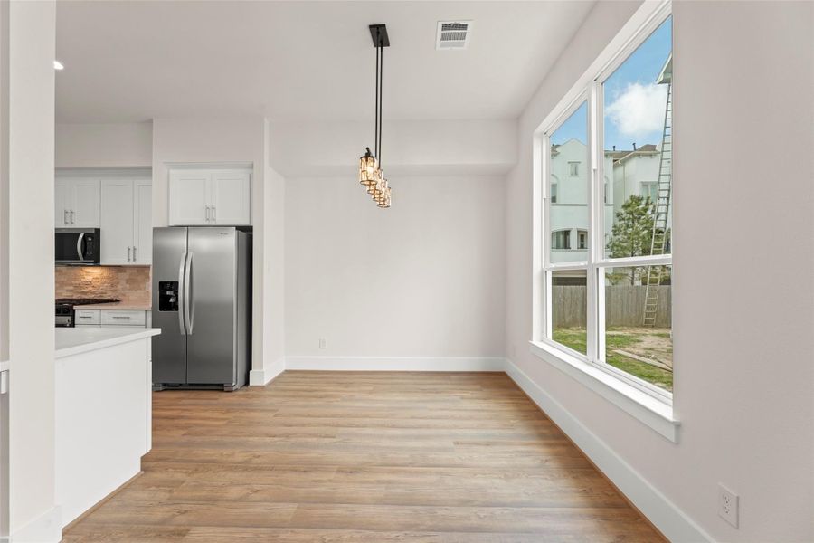 Bright dining area with large windows and modern lighting, adjacent to a sleek kitchen featuring stainless steel appliances and white cabinetry, all set on stylish flooring.