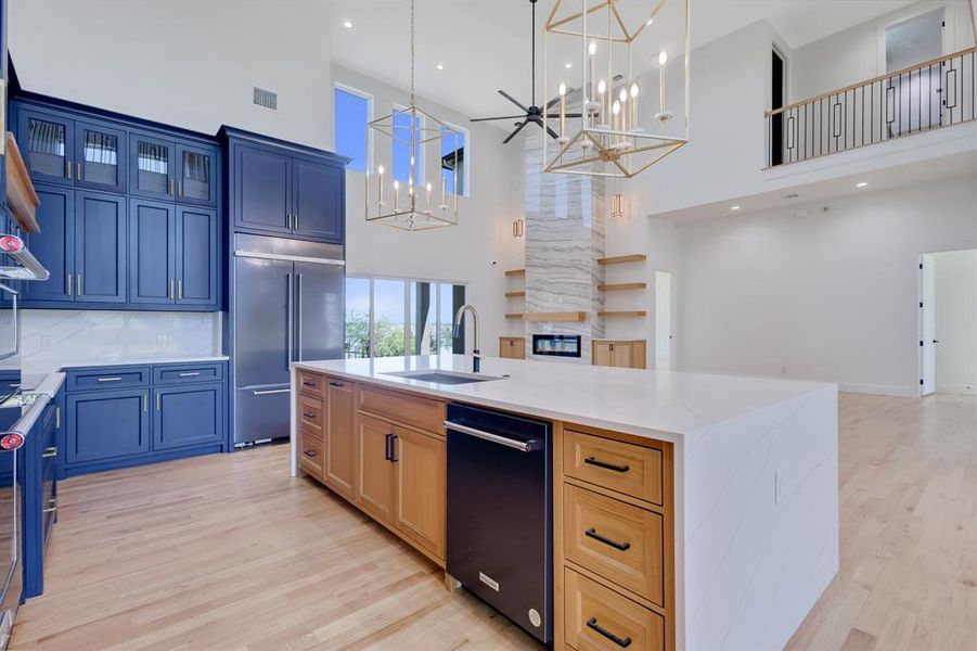 Vaulted ceiling kitchen featuring a large central island with integrated dishwasher, light wood-finish flooring, blue cabinetry with brushed nickel hardware, and stainless steel refrigerator