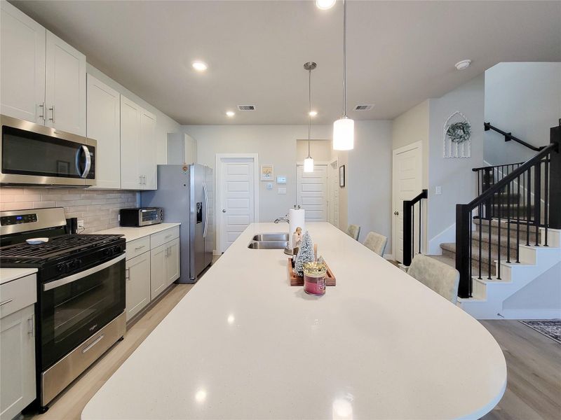 This modern kitchen features sleek white cabinetry, stainless steel appliances, and a spacious island with seating. The open design includes pendant lighting and connects to a stairway, creating an inviting and functional space.