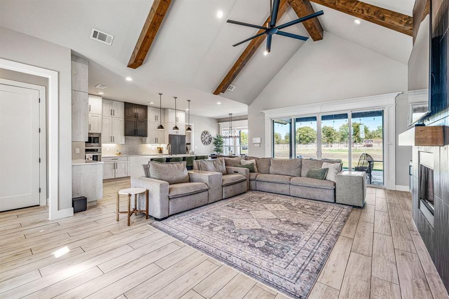 Living room featuring high vaulted ceiling, a ceiling fan, beam ceiling, a glass covered fireplace, and light wood-type flooring