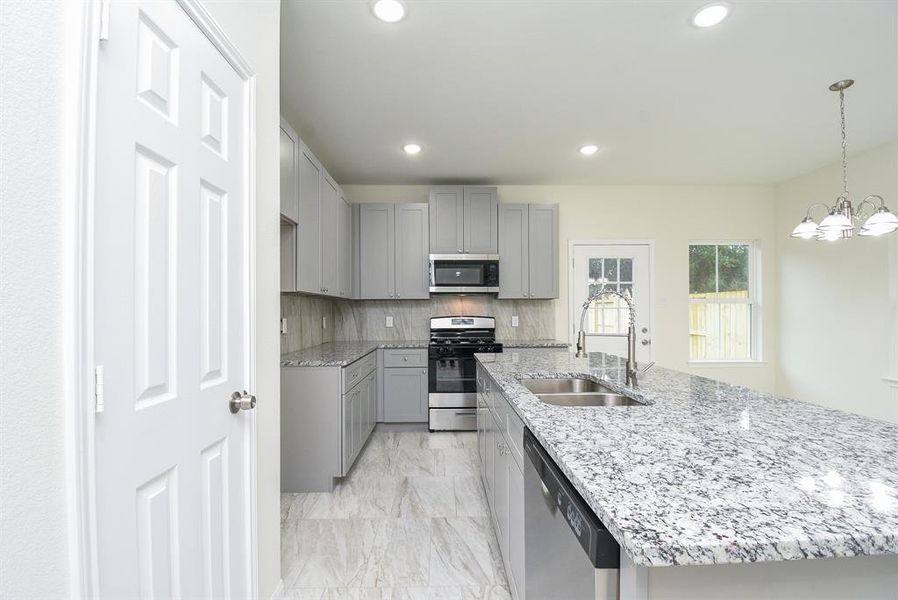 Modern kitchen interior with gray cabinetry, stainless steel appliances, marble countertops, and a chandelier, viewed from a doorway.