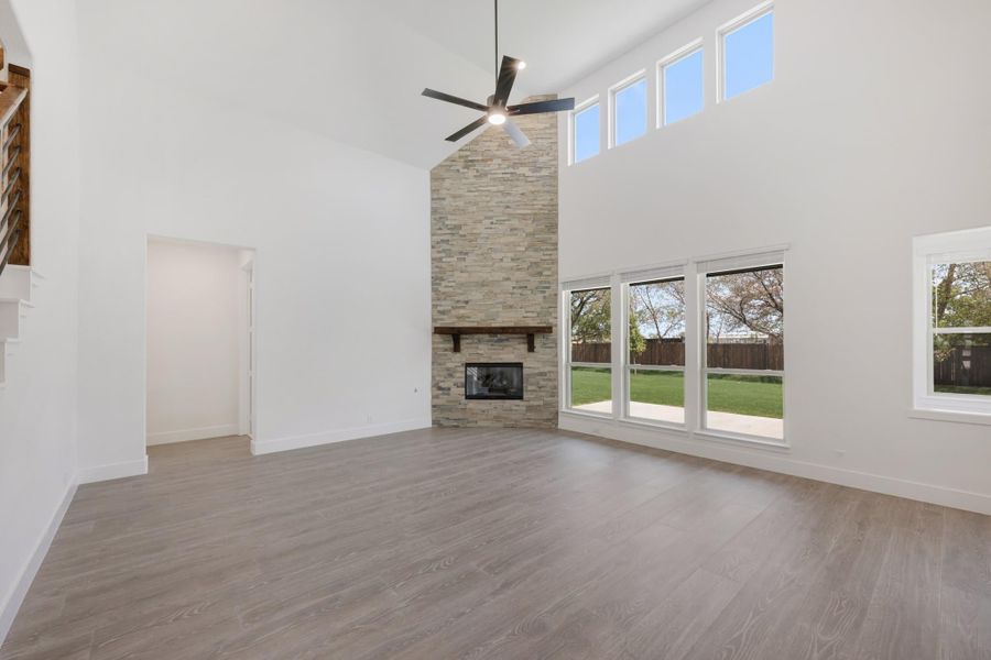 Open living room with vaulted ceiling, stone fireplace, ceiling fan, and large windows overlooking backyard