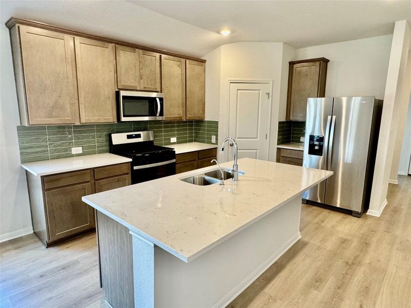 Kitchen featuring stainless steel appliances, decorative backsplash, a kitchen island with sink, light wood finished floors, and light stone counters