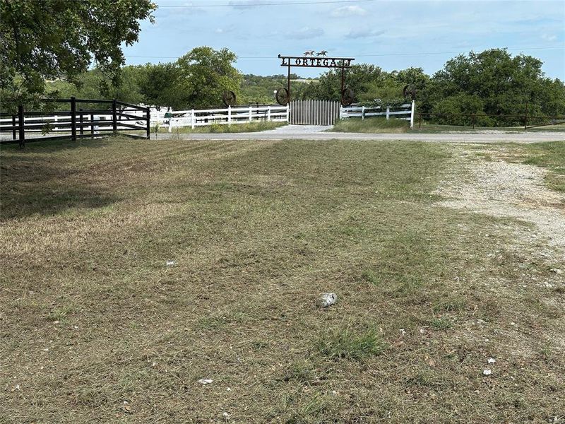Gated entrance featuring a decorative metal archway, white perimeter fencing, and established trees Gated entrance featuring a decorative metal archway, white perimeter fencing, and established trees