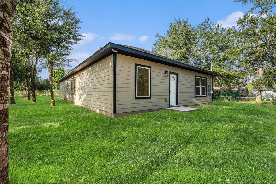This photo shows a single-story home with beige siding and a black roof, surrounded by a spacious, well-maintained lawn and trees, offering a peaceful and private setting.