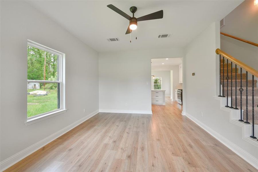 This is a bright, open space featuring light floors, fresh neutral wall paint, and a view of greenery through a large window. There's an elegant staircase with wooden handrails and iron spindles, and the room flows into an adjoining area, suggesting an open floor plan. A ceiling fan provides comfort and a modern touch.