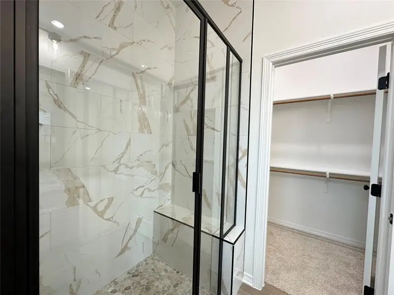Walk-in shower featuring a frameless glass enclosure with matte black trim, large format tile walls with natural veining, a built-in shower bench, and pebble-style flooring