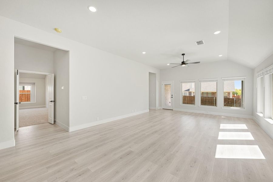 Open living room with vaulted ceiling, ceiling fan, light wood floors, and large windows overlooking backyard