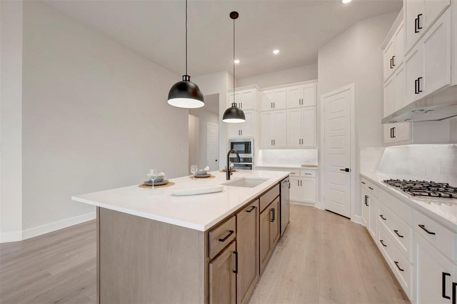 Dual tone kitchen featuring a kitchen island with sink, two tone cabinets, light wood finished floors, pendant lighting, and light stone countertops