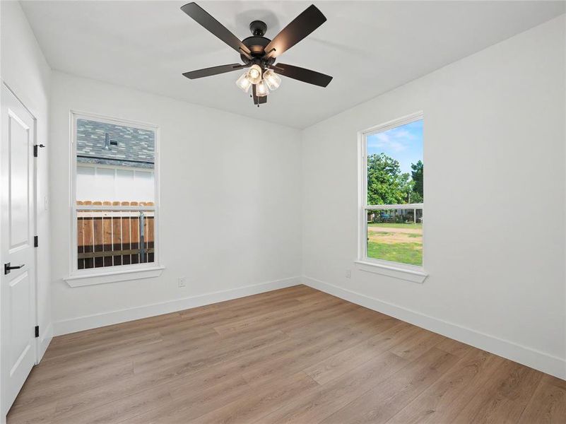 Bright room featuring wood-finish flooring, two large windows, a ceiling fan with light fixture, white walls, and white baseboards Bright room featuring wood-finish flooring, two large windows, a ceiling fan with light fixture, white walls, and white baseboards