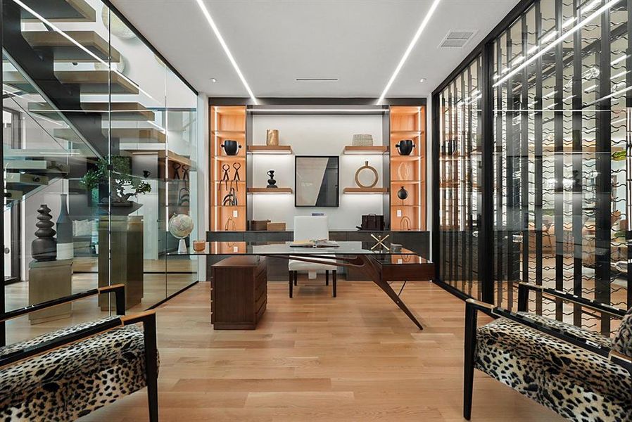 Open-concept office space featuring light wood-finish flooring, recessed linear lighting, built-in shelving with integrated illumination, a glass-enclosed wine storage system, and a glass partition wall