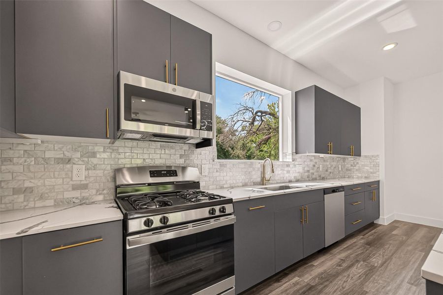 Modern kitchen with sleek gray cabinets and brass handles, featuring stainless steel appliances and a gas stove. The large window provides ample natural light, complementing the elegant marble backsplash and vinyl flooring. Modern kitchen with sleek gray cabinets and brass handles, featuring stainless steel appliances and a gas stove. The large window provides ample natural light, complementing the elegant marble backsplash and vinyl flooring.