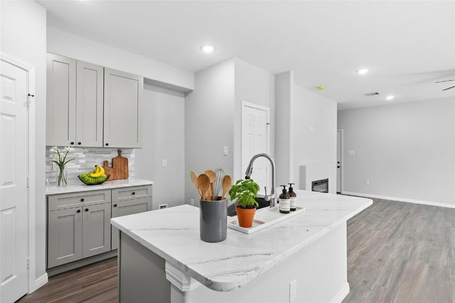 This modern kitchen features sleek gray cabinets, a spacious white marble island with a built-in sink, and contemporary fixtures. It opens to a bright, open-plan living area with luxury vinyl plank flooring, creating a welcoming and functional space. **Photo has been staged.**