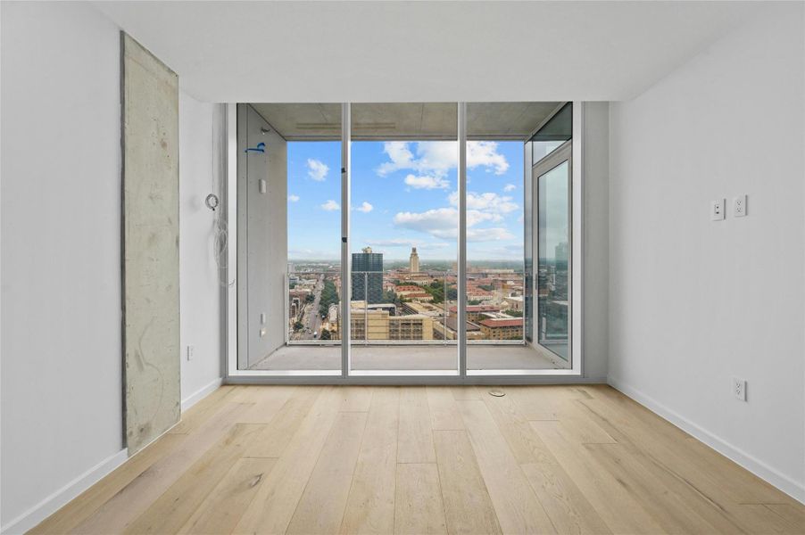 Step into this sleek and contemporary bedroom featuring floor-to-ceiling windows that frame spectacular views of the city and beyond. The natural light floods the room, enhancing the airy ambiance created by the light wood flooring and neutral color palette. The exposed concrete accent wall adds an industrial touch to the minimalist design.